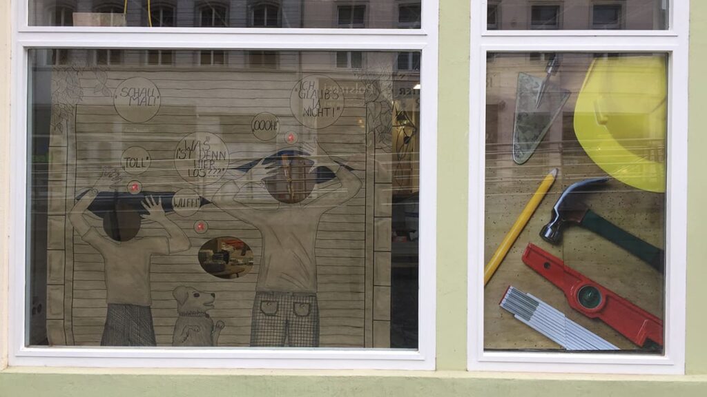 Zwei Schaufenster mit zwei Personen, einem Hund und Sprechblasen im ersten breiten Schaufenster sowie Werkzeug und einem Helm im zweiten schmalen Schaufenster