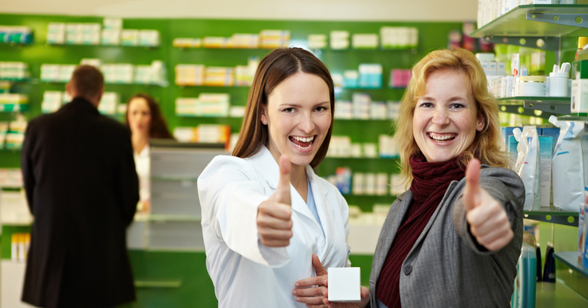 2 Frauen stehen in einer Apotheke und zeigen mit Ihrem Daumen nach oben.