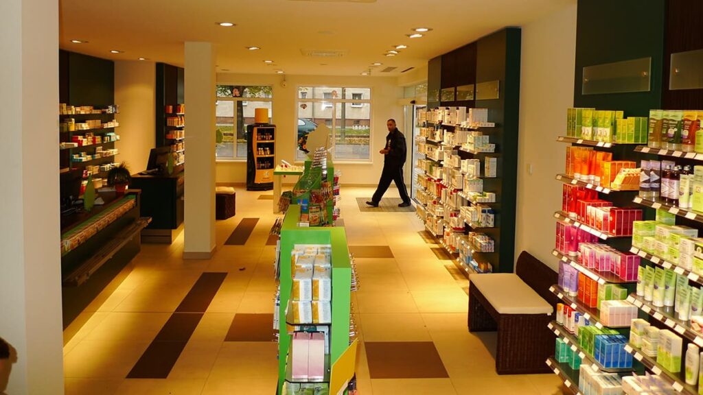 Beleuchteter Verkaufsraum einer Apotheke mit Regalen rechts, dem Kassenbereich links und einem Kunden, der gerade hereinkommt